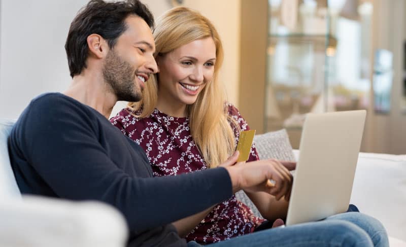 a couple sitting on the couch looking at their laptop and smiling
